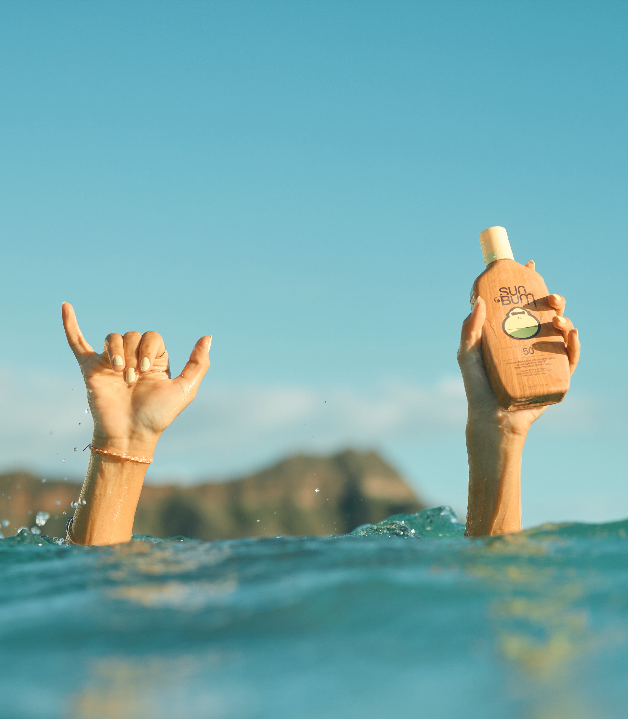 Hands raised from the ocean, one holding SPF 50 lotion, showcasing the Sunscreen Collection – Sun Bum.