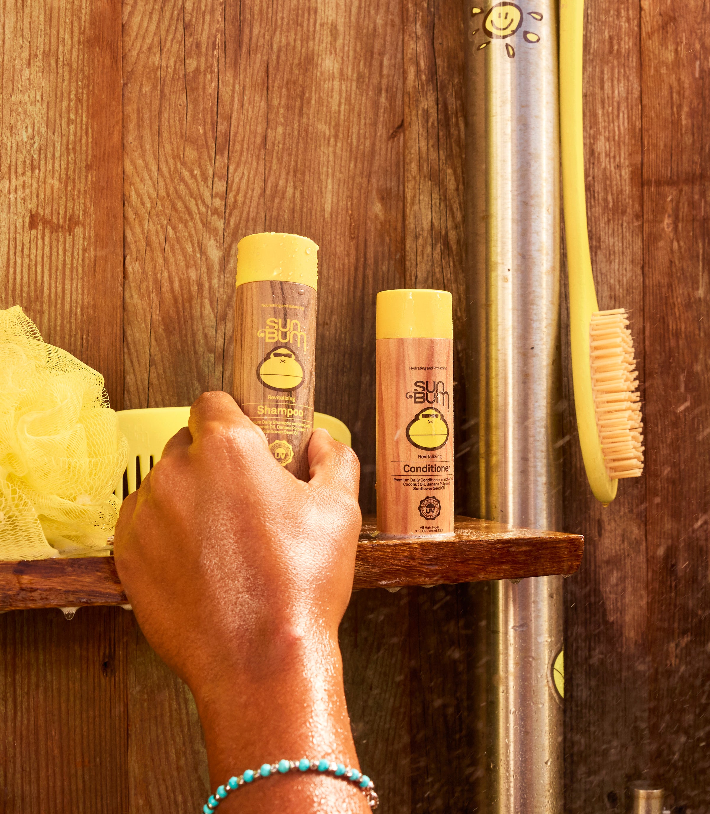 Wet hand reaching for mini Sun Bum shampoo and conditioner bottles on a wooden shower shelf – Travel Hair Care Collection - Sun Bum.
