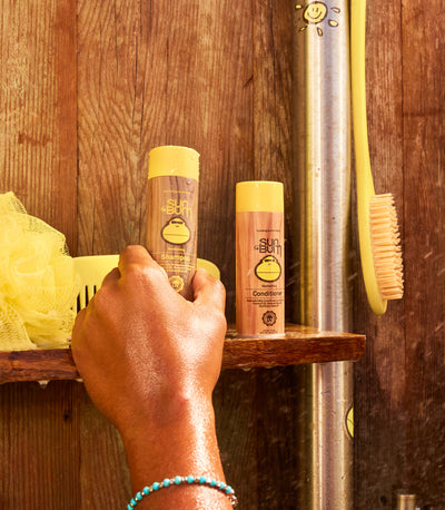 Wet hand reaching for mini Sun Bum shampoo and conditioner bottles on a wooden shower shelf – Travel Hair Care Collection - Sun Bum.