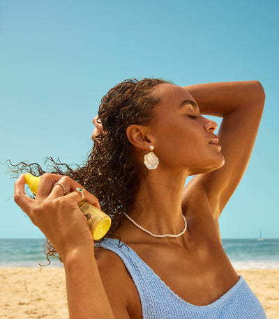 Woman spraying Sun Bum hair treatment on beach with breeze and sunlight – Treatments Collection - Sun Bum.