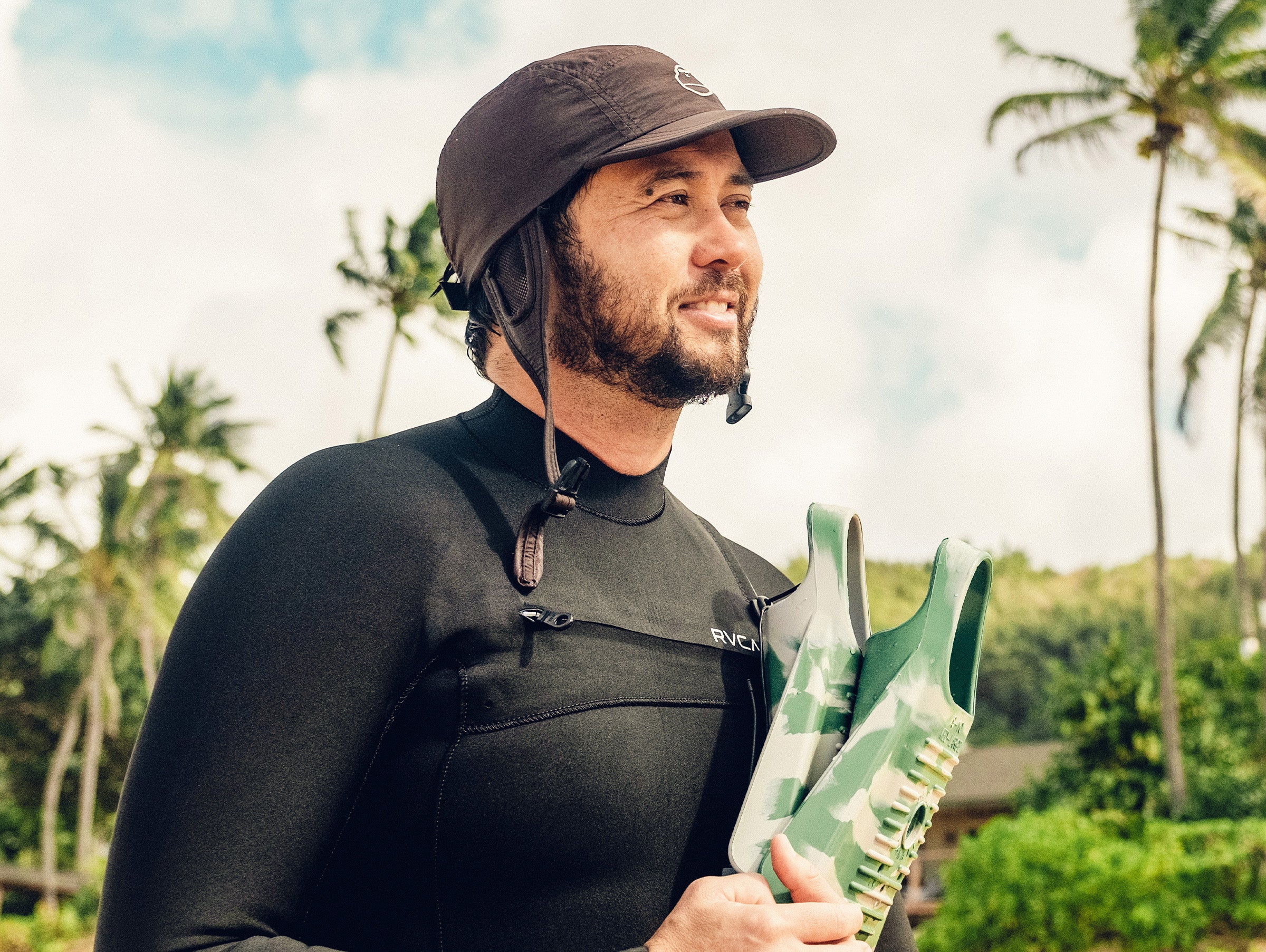 Smiling surfer in a wetsuit holding fins on a tropical beach, ready to hit the waves – Ambassador Collection - Sun Bum