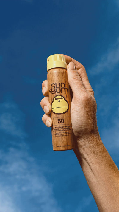 Person holding Sun Bum mini original 5p SPF spray on a blue background