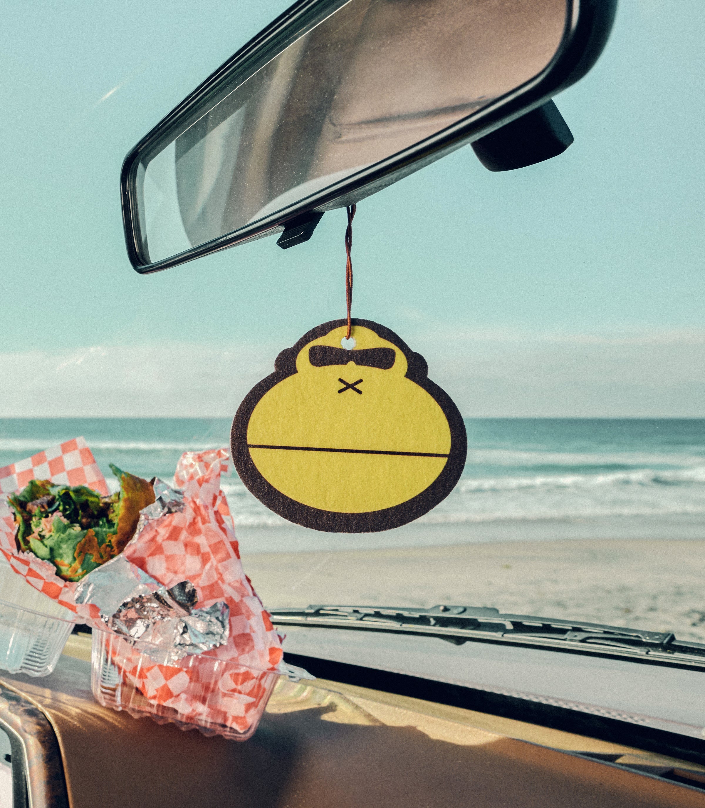 Car air freshener with a cartoon design hanging from a car's rearview mirror with a beach in the background.