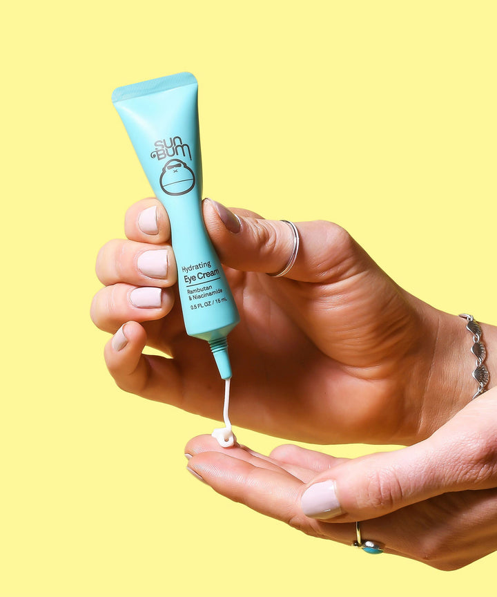A hand squeezing a tube of Sun Bum Hydrating Eye Cream, highlighting its Vitamin C enriched formula with Rambutan and Niacinamide to revive tired, puffy eyes and reduce dark circles. Sun Bum