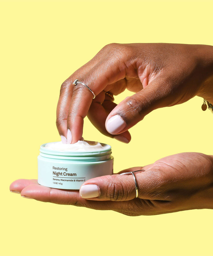 A hand scoops out smooth, creamy Restoring Night Cream from a light green jar, enriched with antioxidants like Banana, Niacinamide, and Vitamin E, to nourish and replenish your skin while you sleep, Sun Bum.