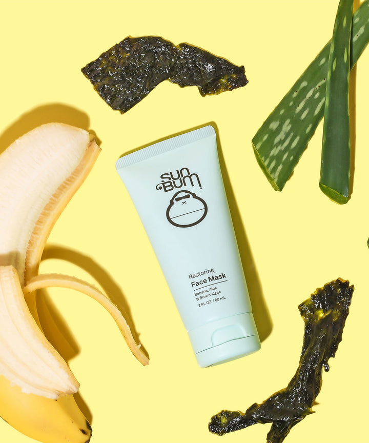 Tube of Sun Bum Restoring Face Mask surrounded by banana, aloe slices, and brown algae on a yellow background, Sun Bum.