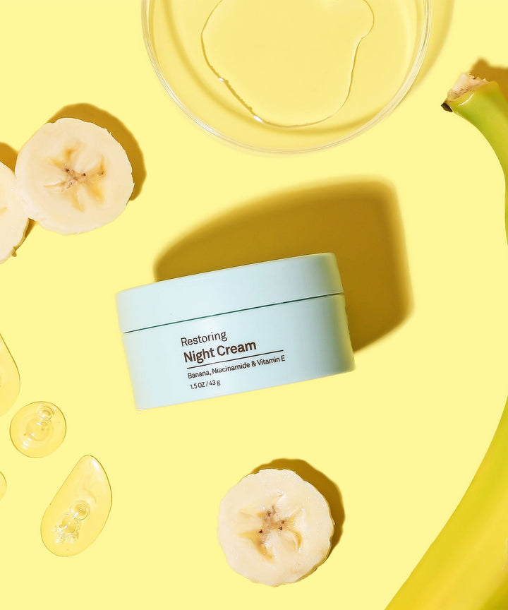 Light blue jar of Restoring Night Cream on a yellow background with banana slices and oil capsules, by Sun Bum.