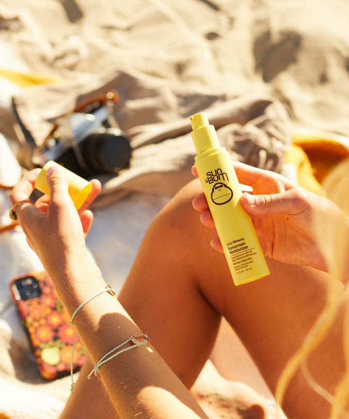 Person applying Sun Bum Daily Mineral Sunscreen Moisturizer SPF 30 at the beach, showing a yellow bottle with lightweight, zinc-based formula perfect for daily skincare protection.