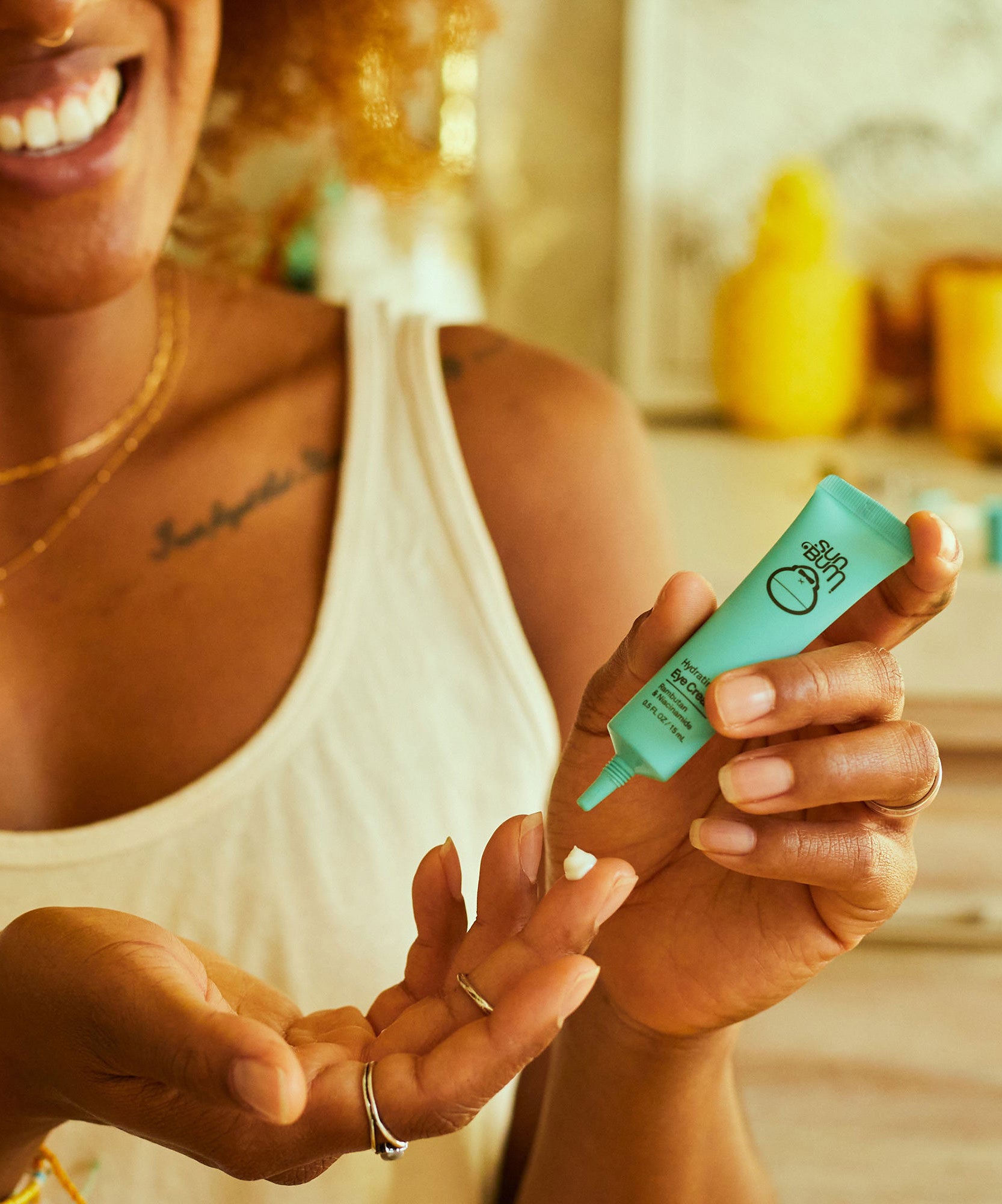 Woman applying hydrating eye cream to hand with Sun Bum product packaging visible in a cozy bathroom setting.