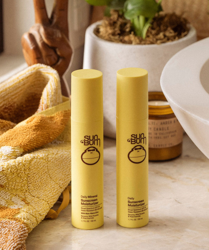 Two bottles of Sun Bum Daily Mineral Sunscreen Moisturizer SPF 30 on a bathroom counter with a towel and a candle in the background. Sun Bum
