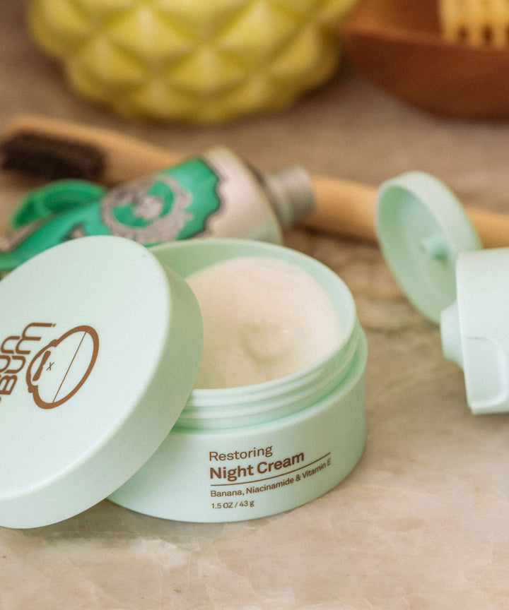 Light blue jar with open lid containing Sun Bum's Restoring Night Cream, enriched with Banana, Niacinamide, and Vitamin E, on a marble countertop.