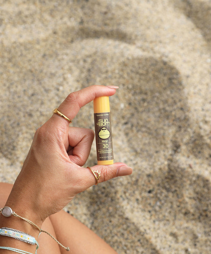 Original SPF 30 Sunscreen Lip Balm - Mango held in hand on the beach, enriched with Aloe and Vitamin E for all-day protection and moisture from Sun Bum.