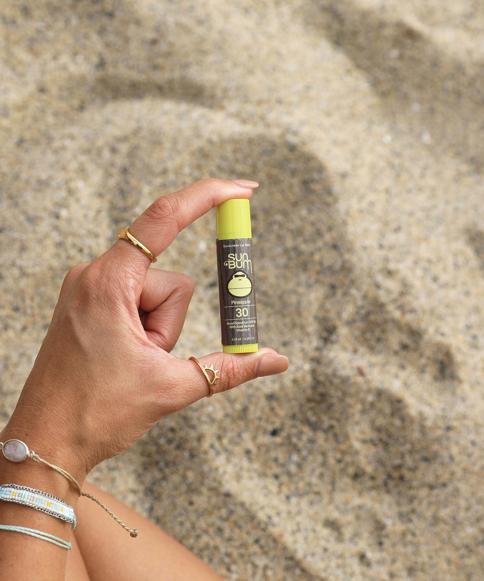 Hand holding Original SPF 30 Sunscreen Lip Balm - Pineapple in a sunny beach setting, Sun Bum